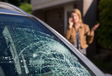 A broken car windshield is in the foreground while an upset looking woman talks on the phone in the background.