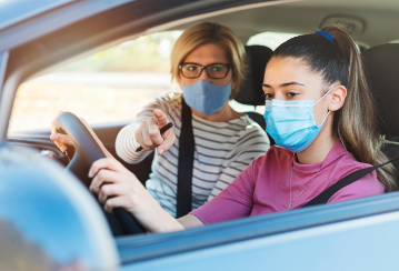 A young woman driver focuses as a driving instructor points and gives instruction. Both are wearing medical face masks.