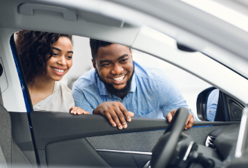 A smiling couple peer into a car through an open driver's side window while considering buying the car.