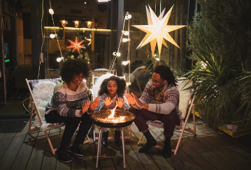 Two parents and their child warming their hands over a portable fire pit in their backyard, with lights and decorations in the background