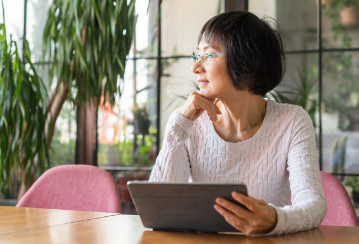 A woman sits at a table holding her tablet and stares off into the distance.