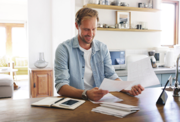 Man looks at papers while sitting at his kitchen table, with more papers and a notebook sitting on the table.