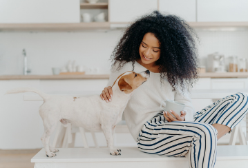 Woman sits at her table and happily pets her dog in her kitchen while enjoying a morning coffee.